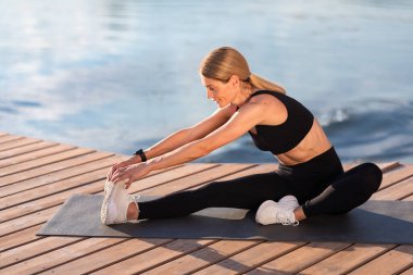 Smiling Athletic Middle Aged Woman Stretching Leg Muscles During Training Outdoors, Sporty Flexible Woman Exercising On Wooden Pier Near River, Making Fitness Workout Outside, Copy Space