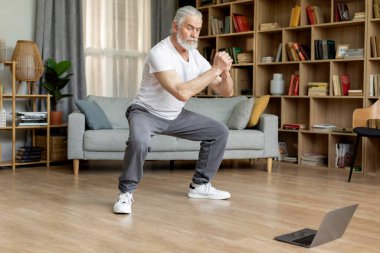 Sporty elderly man in sportswear training at home, watching fitness video on laptop, living room interior, copy space, full length shot. Online fitness class, sports lifestyle for seniors concept