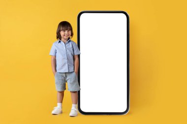Mobile application ad. Cute happy little boy smiling to camera, posing near big blank smartphone with hands in pockets, yellow studio background, empty space