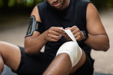 Cropped of black athlete sitting on the ground, wrapping injured knee with elastic bandage, got trauma while exercising outdoors. Injuries during physical activity concept