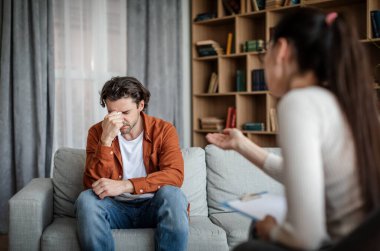 Serious caucasian young lady psychologist calm crying european guy patient in clinic interior. Health care, mental problems, professional medical help, treatment of depression and meeting with doctor