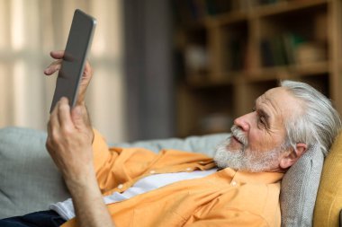 Positive attractive senior man reclining on couch at home, using modern digital tablet, checking social media while chilling alone at weekend, looking at screen and smiling. Leisure and gadgets