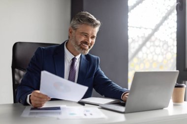 Successful middle aged entrepreneur working with laptop and papers at workplace in modern office, smiling mature businessman in suit sitting at desk and managing financial reports, copy space