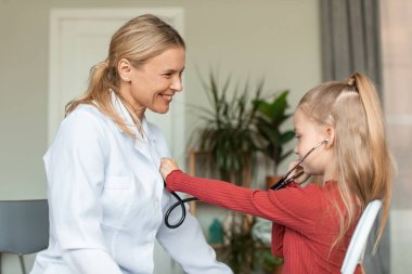 Happy little girl visiting pediatrician and playing with doctor, listening docs heart beating with stethoscope, sitting in clinic and smiling to each other