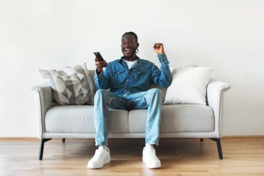 Excited African American Man Watching TV Shaking Fists In Joy Holding Remote Controller Sitting On Sofa At Home. Television Programming, Victory Celebration Concept