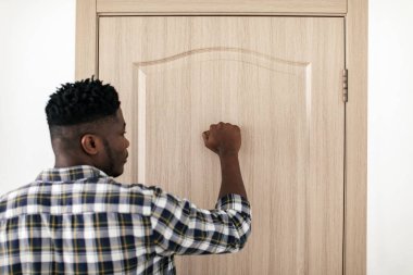 Rear View Of Black Guy Knocking At Closed Entry Door Of Apartment Standing In Doorway Indoors. Real Estate Offer And Business Concept. Selective Focus On Fist