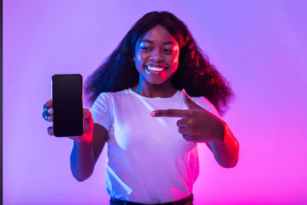 Smiling millennial black female pointing at cellphone with empty screen in neon light, mockup for website or mobile app, selective focus. Cellphone touchscreen template with blank space for ad