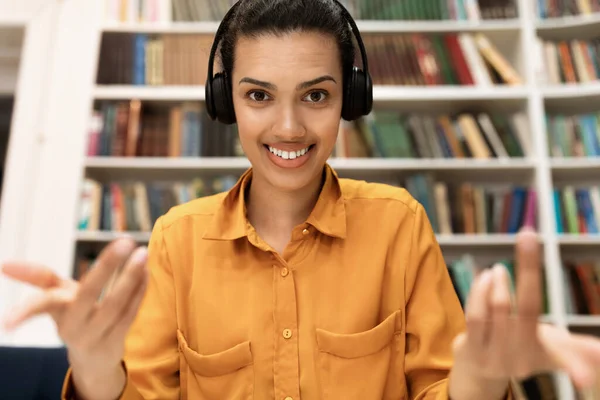 Online tutoring concept. Excited female teacher having video call, talking and gesturing at camera during online lesson, sitting in library interior