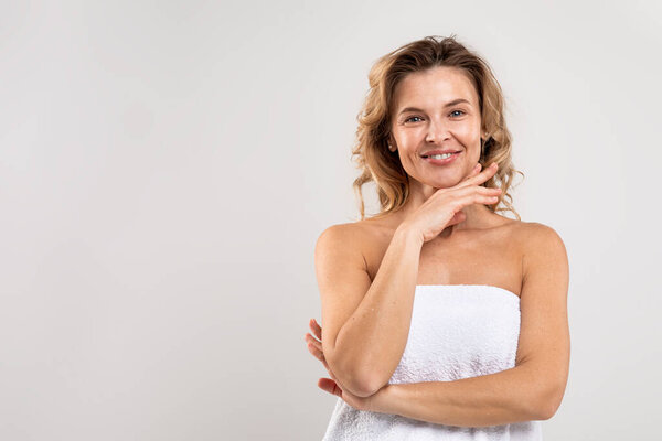 Female Beauty. Attractive Middle Aged Woman Wrapped In Bath Towel Posing In Studio, Happy Good-Looking Lady With Beautiful Skin Smiling At Camera, Standing Over Light Grey Background, Copy Space