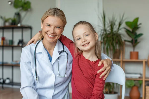 Portrait of friendly female doctor embracing with little cute girl patient in hospital, looking and smiling togethe at camera. Good children healthcare concept