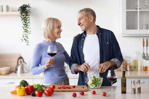 Loving senior couple celebrating marriage anniversary, happy mature man and woman drinking red wine, having conversation while making dinner together, white kitchen interior, copy space