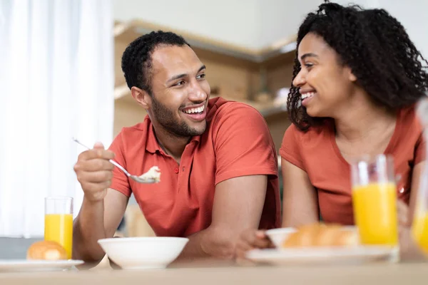 Laughing young african american wife and husband in same t-shirts have breakfast and enjoy communication at free time in kitchen interior. Human emotions, proper nutrition, love and relationships