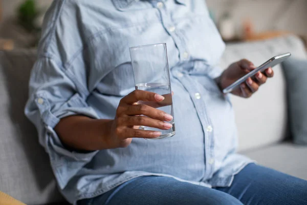 Unrecognizable Pregnant Black Female Using Smartphone Holding Glass Water Sitting — Foto de Stock