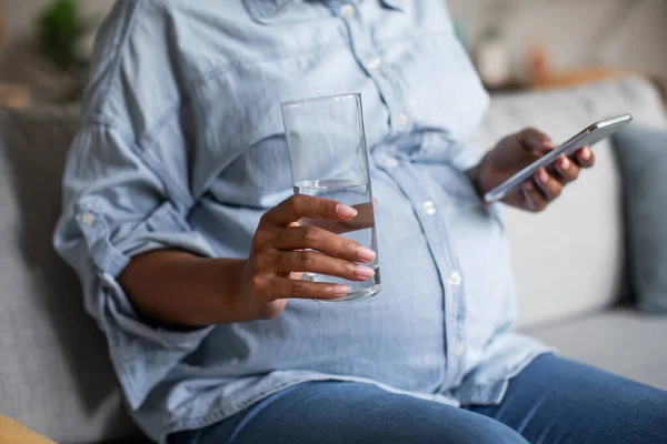 Unrecognizable Pregnant Black Female Using Smartphone Holding Glass Water Sitting — стоковое фото