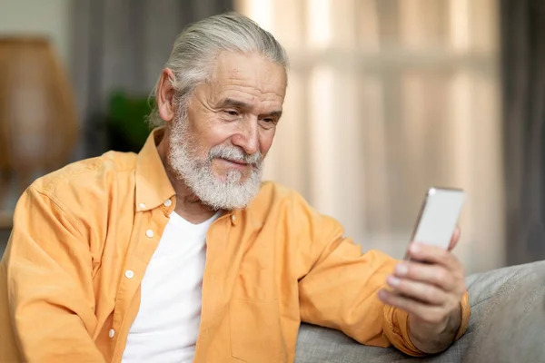 Closeup of positive attractive old man in casual outfit sitting on sofa in living room, using brand new mobile phone at home, having online party with his friends or family, copy space