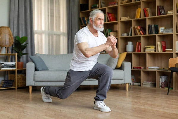 Motivated athletic senior man in sportswear and sneakers doing lunges at home, looking at copy space. Good-looking grandfather exercising in living room while coronavirus pandemic, full length shot