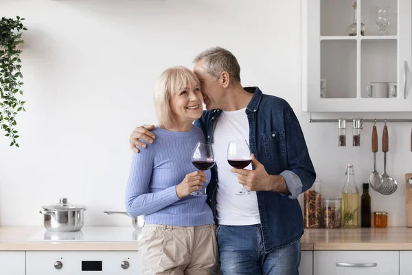 Romantic senior couple drinking red wine at kitchen, happy elderly man and woman in casual outfits standing by kitchen table with glasses, hugging and cuddling, enjoying time together, copy space