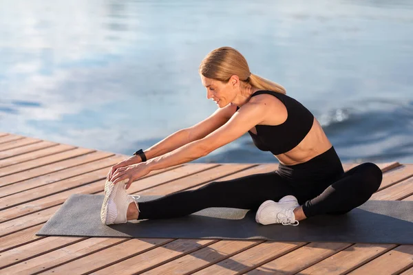 Smiling Athletic Middle Aged Woman Stretching Leg Muscles During Training Outdoors, Sporty Flexible Woman Exercising On Wooden Pier Near River, Making Fitness Workout Outside, Copy Space