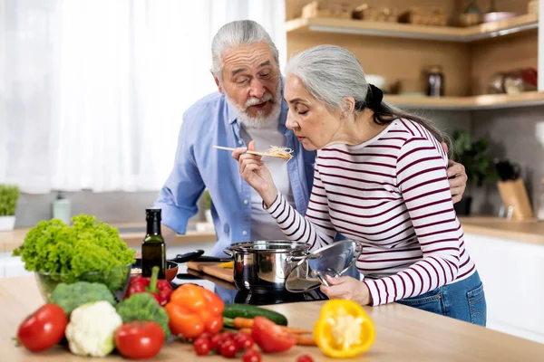Beautiful Senior Woman Tasting Food While Cooking With Husband In Kitchen Together, Happy Elderly Spouses Making Delicious Meal For Lunch, Preparing Spaghetti With Vegetables At Home, Copy Space