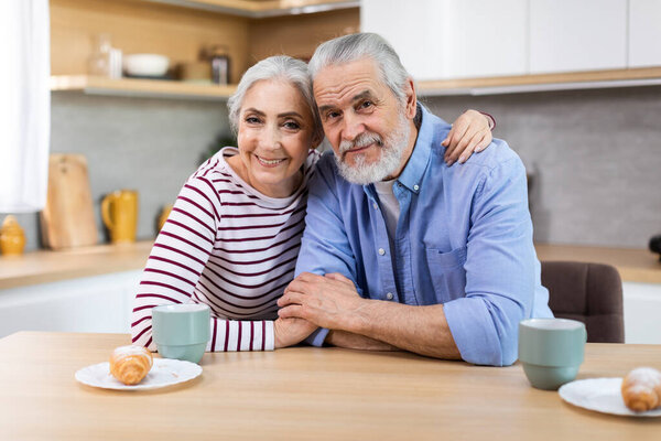 Portrait Of Happy Senior Spouses Posing At Table In Kitchen, Cheerful Elderly Couple Having Coffee With Croissants And Smiling At Camera, Enjoying Spending Time Together At Home, Closeup Shot