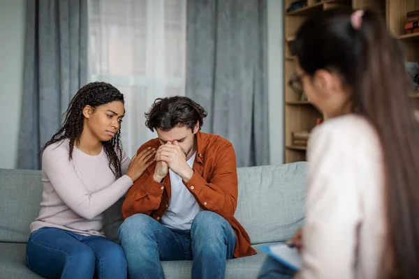 Sad millennial black lady calms european man at meeting with psychologist in office clinic interior, copy space. Family psychological therapy, grief, tragedy, depression and illness treatment together