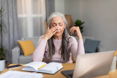 Sad tired caucasian elderly gray-haired woman takes off her glasses, wipes eyes in living room interior. Overwork, vision problems, work at home, business with technology during covid-19 quarantine