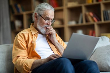 Pensive grandfather stylish grey-haired senior man websurfing while resting at home, sitting on couch and using modern laptop, touching his chin, reading blog, living room interior, copy space