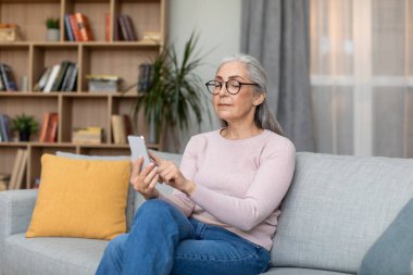 Serious concentrated caucasian elderly gray-haired lady in glasses typing on phone, use new app in living room interior, free space. Lifestyle, modern device, chat in social networks, ad and offer