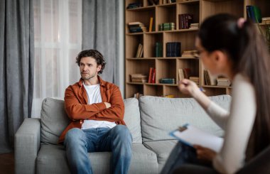 European young woman psychologist talk with dissatisfied guy, patient ignoring doctor advice in clinic interior. Mental problems, professional medical care, fight against depression, help and support