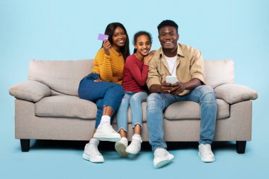 Online shopping concept. Excited black parents and their daughter using cellphone and credit card, purchasing in internet, sitting on couch over blue studio background