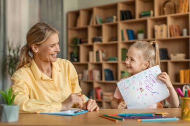 Cute little girl showing attractive woman psychologist her drawing, kid having therapy session with child development specialist at clinic