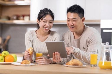 Smiling chinese spouses having breakfast and using modern digital tablet together, sitting at kitchen table, drinking orange juice, eating croissants, surfing on Internet, copy space