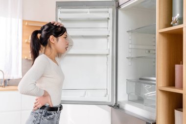Overwhelmed pretty young korean lady checking her fridge at home, desperate hungry woman looking inside empty cooler with no groceries and touching her head, side view, copy space
