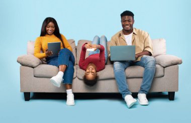 Gadgets addiction concept. African american family of three using different electronic devices, sitting on sofa on blue background. Parents and their daughter with modern gadgets