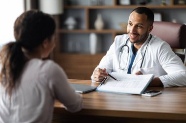 Friendly handsome african american young man doctor having conversation with female patient, practitioner showing treatment plan and smiling, giving details, modern clinic interior