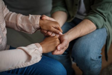 Cropped millennial african american lady doctor calms caucasian unhappy male patient and holding hands in clinic interior, close up. Professional support, medical care, mental problems, depression