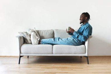 Cheerful African American Millennial Man Drinking Coffee Holding Cup Relaxing Enjoying Cozy Weekend At Home. Guy Sitting On Couch Indoor Concept. Side View Shot