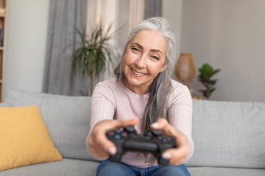 Glad smiling caucasian old gray-haired female with joystick playing online game in living room interior. Leisure time and entertainment at home, fun alone and technology during covid-19 quarantine
