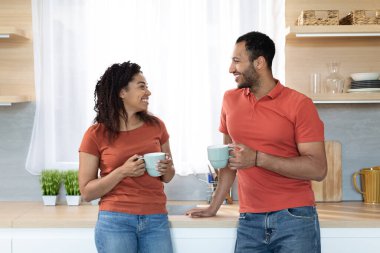 Happy relaxed young african american guy and woman in same red t-shirts with cups enjoy drink and free time together in cozy kitchen interior. Coffee break, couple relationship, love and breakfast