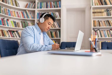 Side View Of Korean Man Working Or Learning Online On Laptop Taking Notes Wearing Headphones Sitting At Desk In Study Room With Bookshelves. E-Learning, Freelance Career Concept