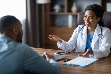 Friendly attractive african american young woman doctor having conversation with male patient, general practitioner consulting young man, gesturing and smiling, giving details, modern clinic interior