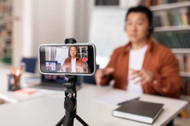 E-Teaching. Japanese Male Tutor Filming Lecture On Smartphone For Educational Blog Having Online Lesson Sitting At Workplace. Modern Education Concept. Selective Focus On Mobile Phone