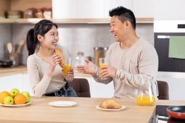 Happy asian couple having snack at kitchen, positive korean man and woman sitting at table, drinking fresh orange juice, eating croissants, having conversation, enjoying time together at home