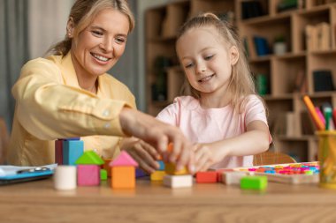 Games and education concept. Happy professional woman teacher exercising with cute girl, playing wooden development blocks at classroom, sitting at desk