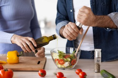Unrecognizable elderly man and woman in casual outfits cooking together at home, making delicious vegetable salad, add olive oil and seasoning, kitchen interior. Healthy diet for seniors