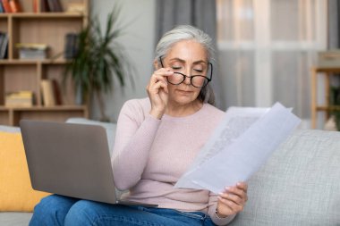 Serious caucasian old gray-haired lady in glasses works with documents and laptop in living room interior. Modern technology, freelance, pay tax, bookkeeping, job remote at home due covid-19 outbreak