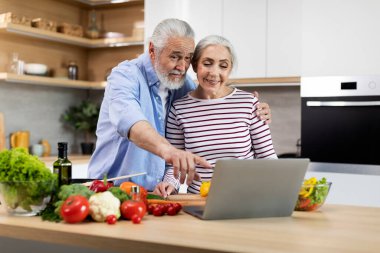 Happy Married Senior Couple Using Laptop While Cooking Healthy Food In Kitchen, Cheerful Elderly Spouses Checking Online Recipe On Computer While Preparing Vegetable Meal At Home, Free Space