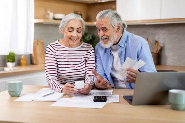 Smiling Elderly Spouses Checking Receipts While Planning Family Budget At Home, Happy Married Senior Couple Counting Monthly Spends, Sitting At Table With Laptop In Kitchen, Closeup Shot