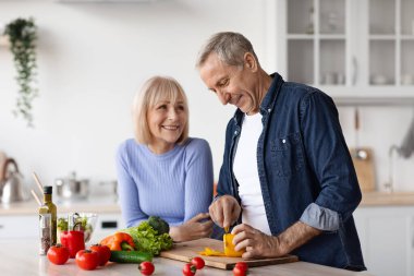 Beautiful senior spouses having conversation and smiling while cooking at home, cheerful pretty elderly woman supporting her husband preparing healthy dinner, cutting vegetables, copy space