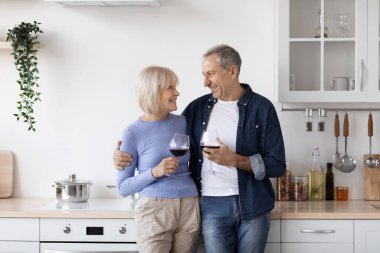 Romantic elderly husband and wife standing by kitchen table, embracing, drinking wine at home, enjoying time together, copy space. Love, family, affection, relationships concept